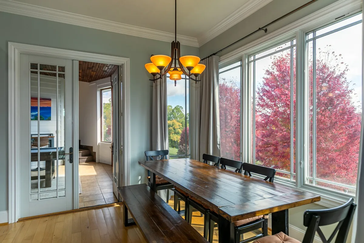 The focus here is on a large dining table, illuminated by a striking chandelier and surrounded by windows with views of the outdoors. The table is set for a group, inviting family and friends to gather for celebrations or everyday meals.