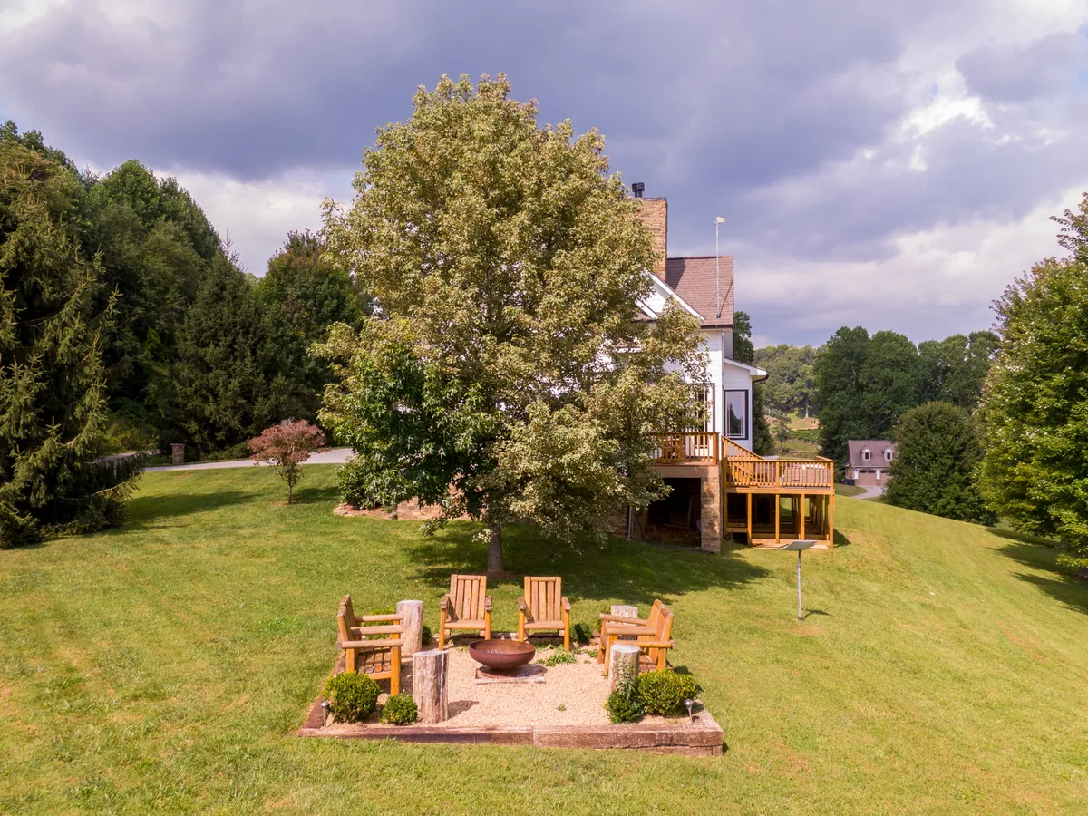 The fire pit area with Adirondack chairs arranged around it sits under a large tree, adjacent to the house's deck and surrounded by lush greenery. The inviting setup is ideal for cozy evenings, storytelling, and enjoying nature with friends or family.