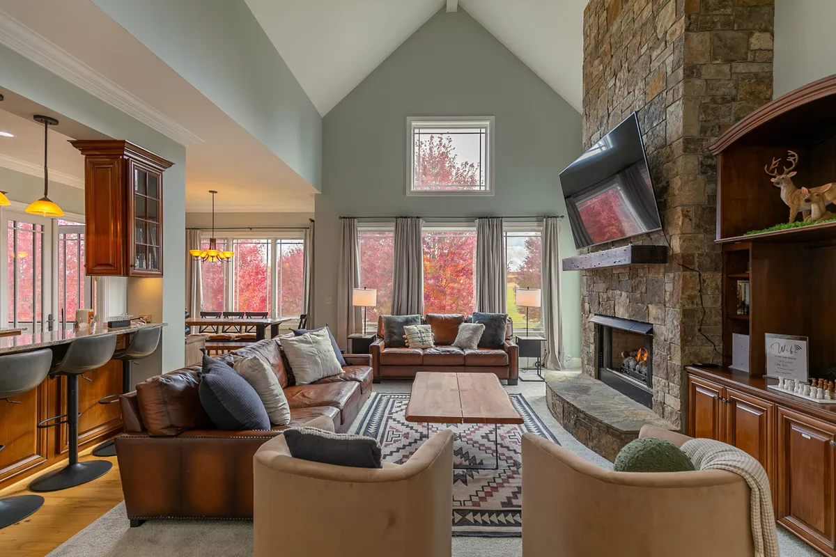 This image showcases the soaring great room with a stunning floor-to-ceiling stone fireplace as the centerpiece. Large windows fill the room with natural light and offer expansive views of the autumn foliage outside. Plush, comfortable seating is arranged around a wooden coffee table, creating an inviting atmosphere for gathering, relaxing, and socializing. The entertainment center suggests options for movie nights or watching television with company.