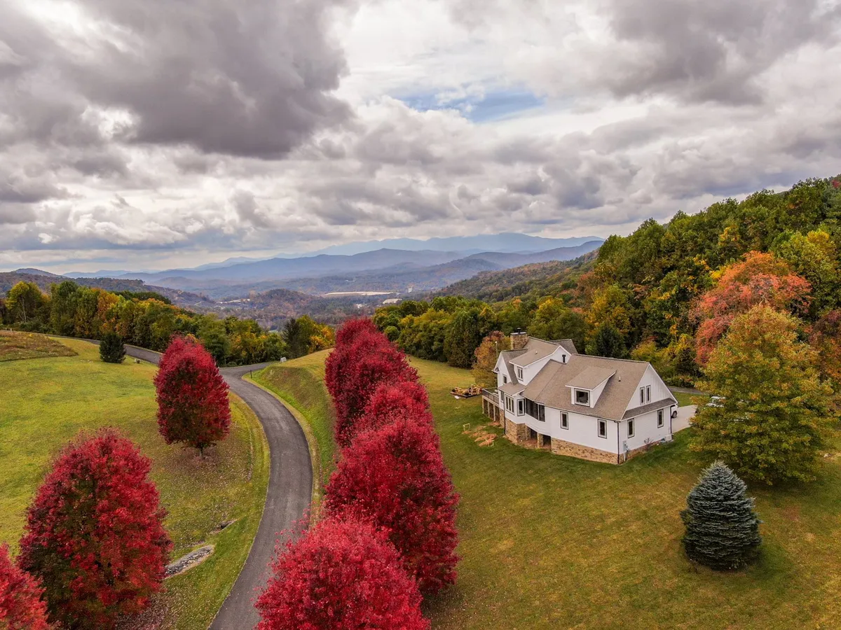 This image presents the house nestled amongst colorful trees, with a winding road leading toward the distant mountain range. The autumn foliage is vibrant, and the overall scene is peaceful and inviting, evoking the scenic beauty of the Blue Ridge Parkway area.