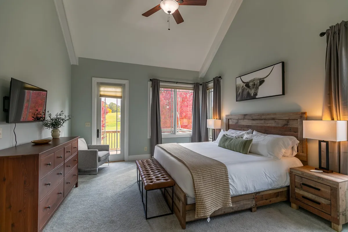 A serene king-sized bedroom featuring a large window with mountain views, a wooden dresser, and tasteful decor, including a whimsical cow artwork. The space is bright, tranquil, and inviting, perfect for relaxation and privacy after an active day.