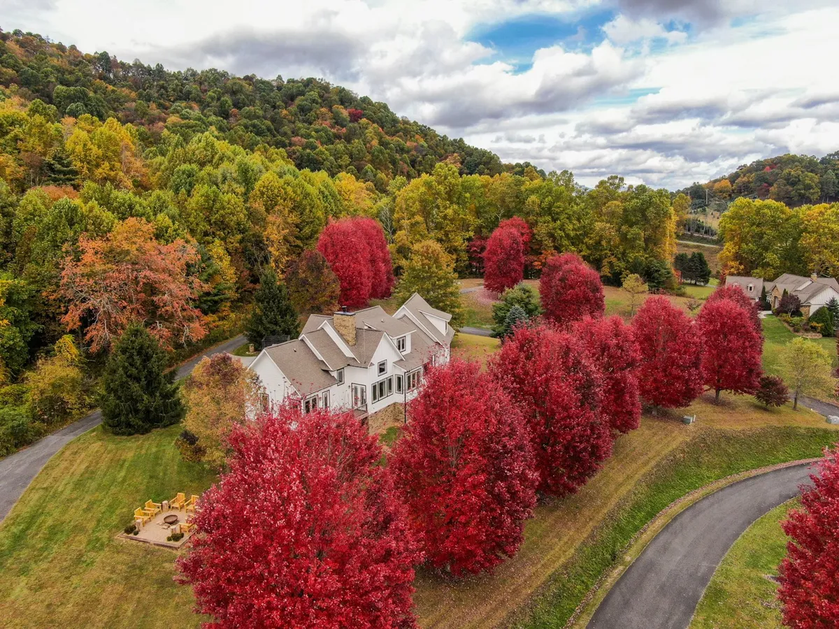 The image showcases a spacious single-family mountain home set against a backdrop of vibrant red autumn trees. A winding driveway leads up to the house, and an inviting outdoor seating area suggests space for relaxation and gatherings. The overall scene is serene and picturesque, emphasizing tranquility, nature, and ample space for group enjoyment.
