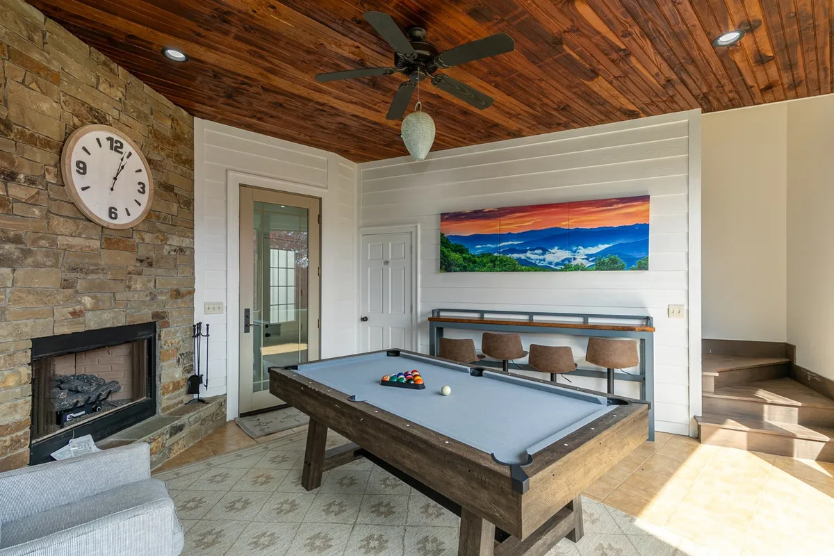 This image features a vibrant three-season room with a pool table at the center, a cozy fireplace along the wall, and a bright, welcoming seating area. Decorative wall art and a clock add character while natural light pours in from windows, creating a perfect space for both game nights and relaxation by the fire.