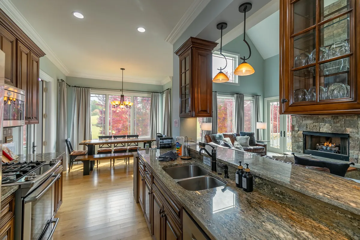 A wide-angle view encompassing the kitchen island, large windows letting in natural light, a spacious dining table, and a cozy fireplace. The scene creates a seamless flow between cooking and dining areas, emphasizing spaces where groups can gather and interact.
