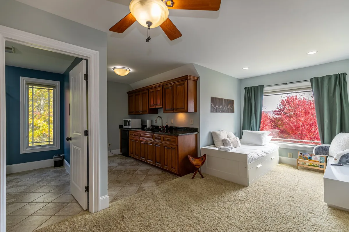An inviting loft area with a daybed adorned with plush cushions, a kitchenette area, and a window overlooking autumn foliage. The space feels warm and perfect for relaxing, reading, or enjoying a quiet snack.