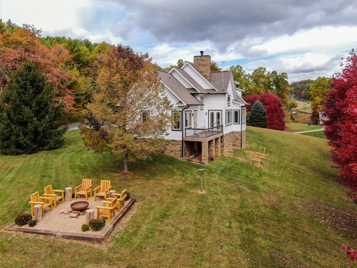 The image features a charming house with a stone base set amidst trees sporting fall foliage. In the foreground, a cozy fire pit area invites guests to gather and relax, suggesting the perfect end to a day of adventure under the stars.