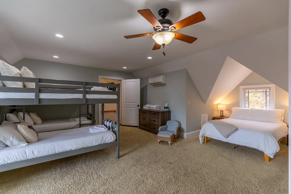 A cozy bedroom with a queen bed, bunk beds, a window letting in natural light, and a ceiling fan. The environment is warm and suitable for families or groups, offering both comfort and versatility for various sleeping needs.